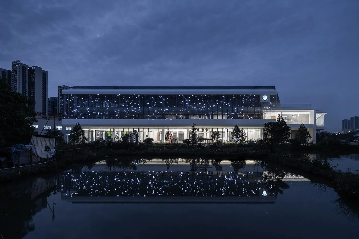 A modern office building reflecting in a pond at dusk, illuminated by numerous small lights.
