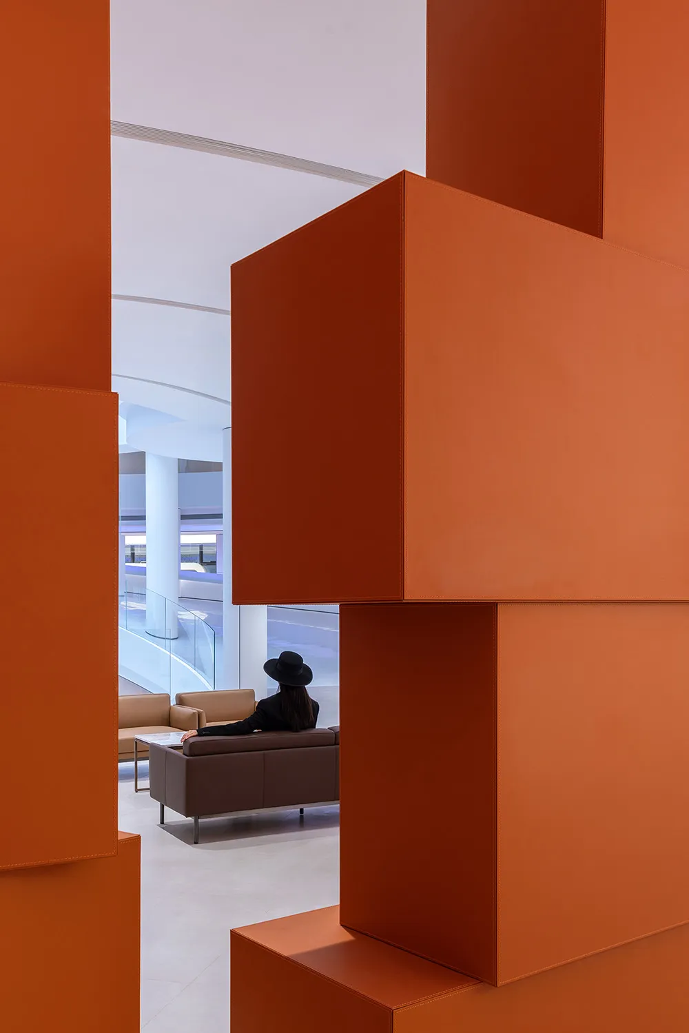 A modern lobby with large, orange, geometric structures and a woman wearing a black hat sitting on a brown sofa.