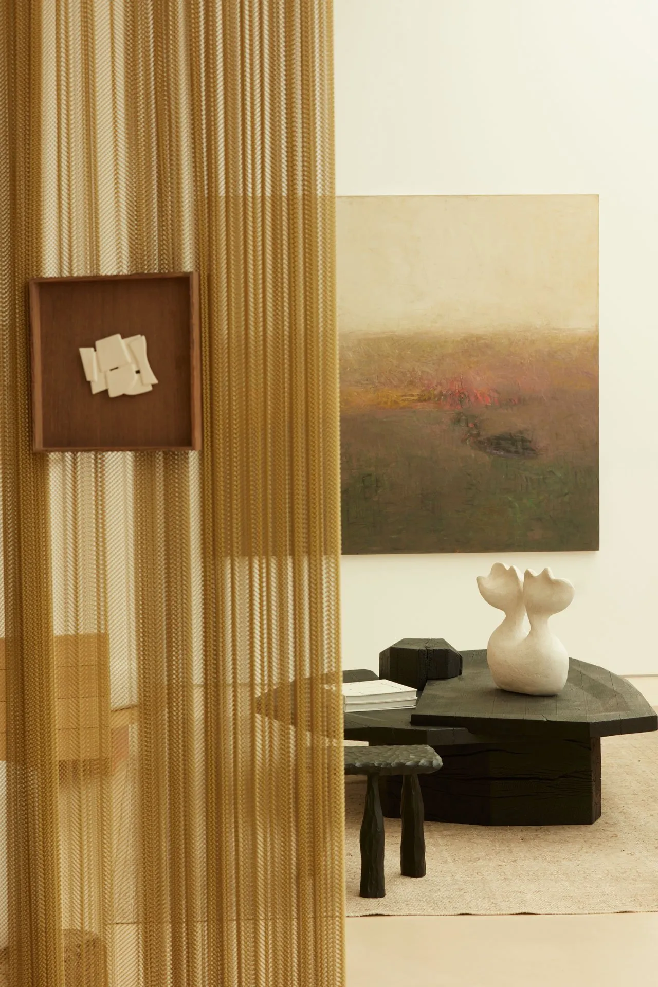 Modern minimalist living room featuring a black wooden coffee table surrounded by textured, woven textiles. Artwork on the wa