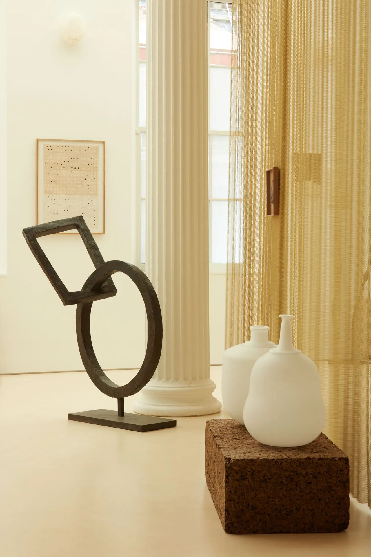 A modern interior featuring an abstract metal sculpture on a pedestal and two white vases on a cork block, set against a yell
