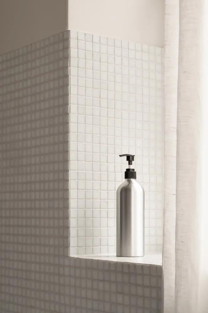 A metallic bottle with a black dispenser cap, placed on a shelf against a backdrop of white square tiles.