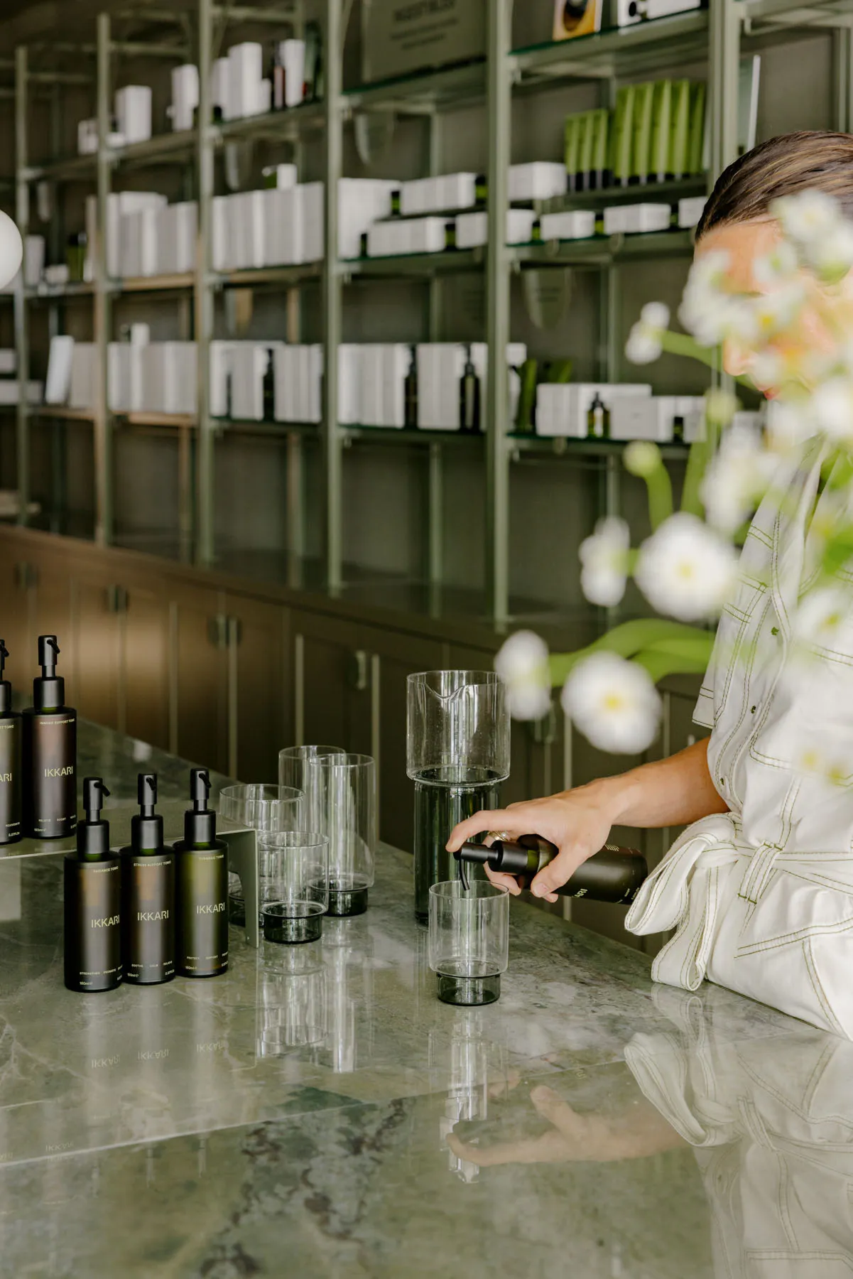 Person pouring a container of liquid into a clear glass in a spa setting with glass shelves and flower arrangements.