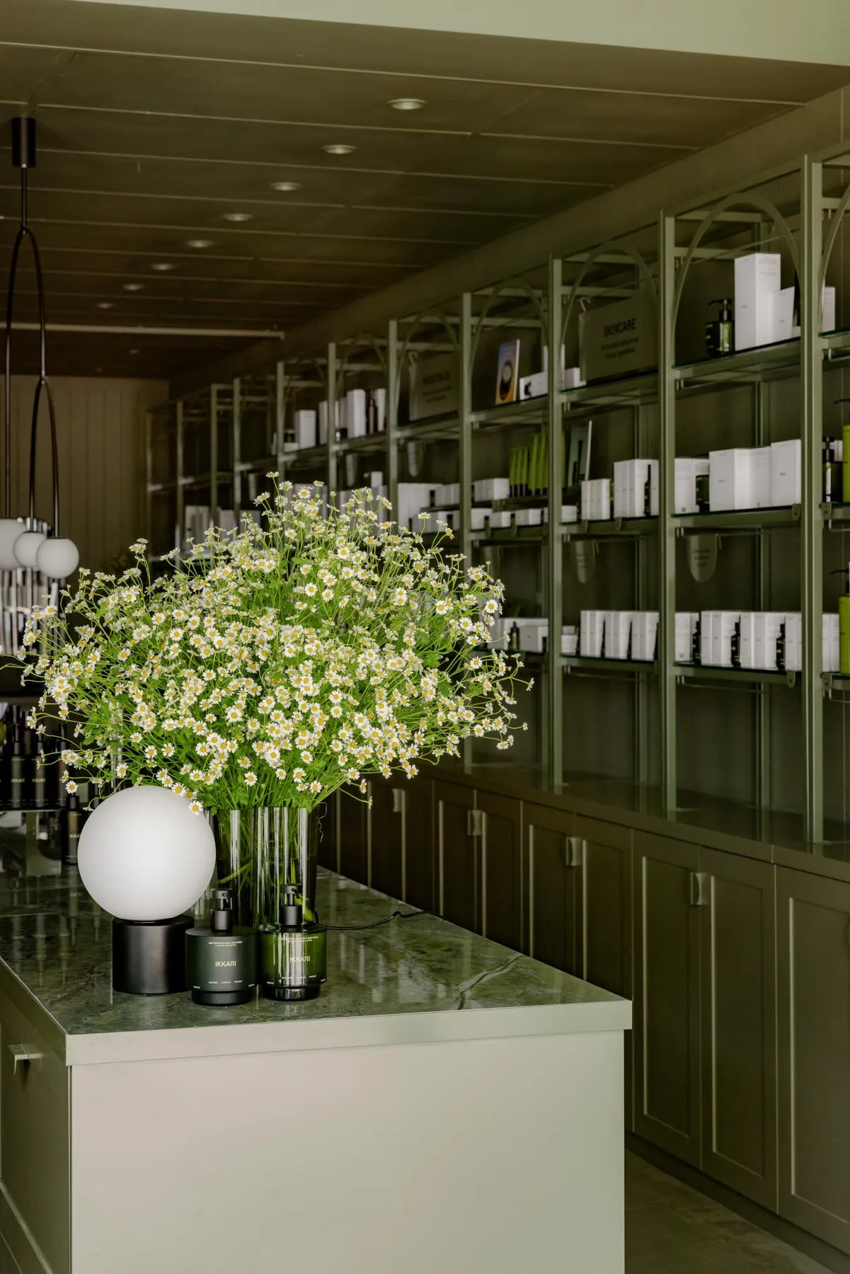 A spa interior with a large bouquet of daisies in a green glass vase, surrounded by shelves filled with skincare products and