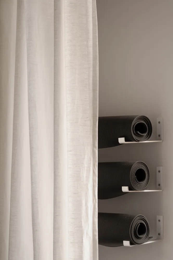 A white curtain with white metal hooks on a white metal rod with three black yoga mats rolled up on the hooks.