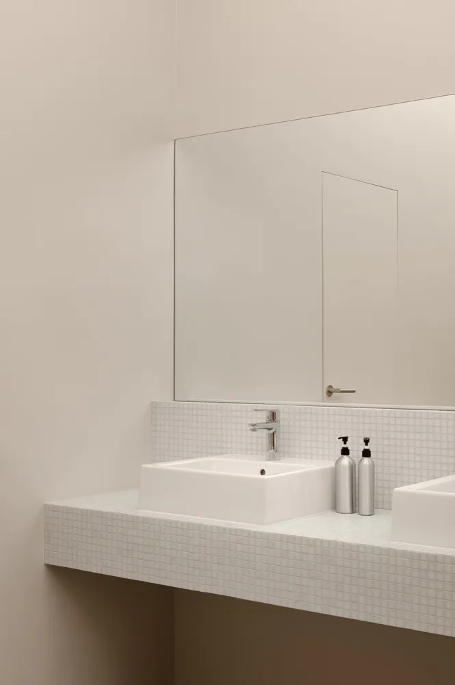 A modern restroom features a white mosaic-tile backsplash with steel trim, a sleek white sink, and silver soap dispensers. Th