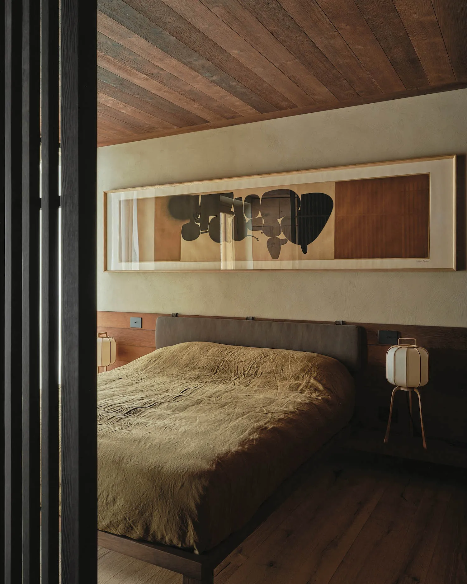 A minimalist bedroom featuring a wooden bed frame and headboard, with a large framed abstract painting above it.