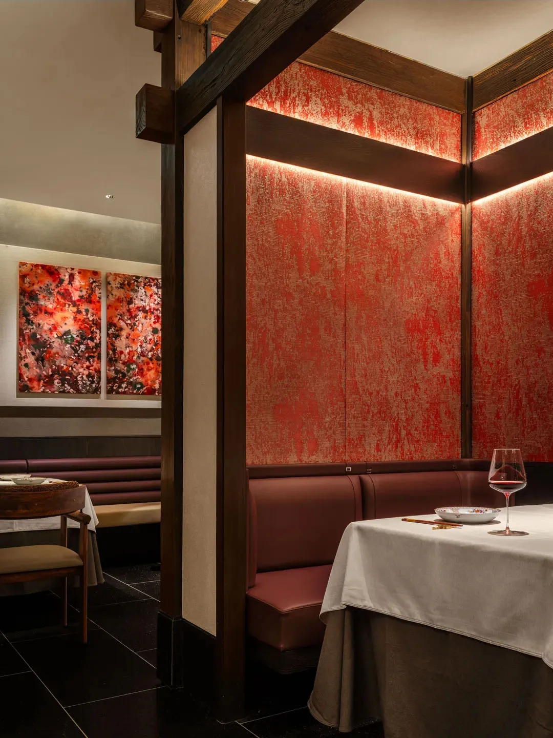 A modern restaurant room with red textured walls, wooden beams, and a booth with a table and wine glass.