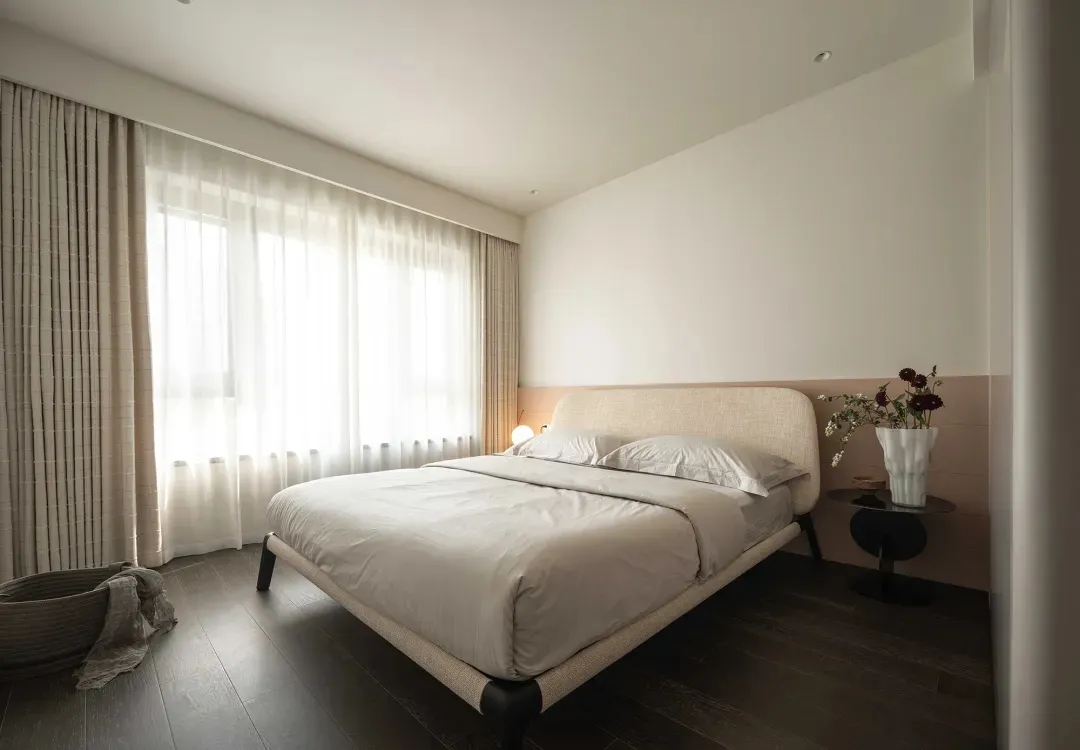Modern bedroom featuring a taupe and wood upholstered bed with a minimalist design, white curtains, and a vase of flowers on 