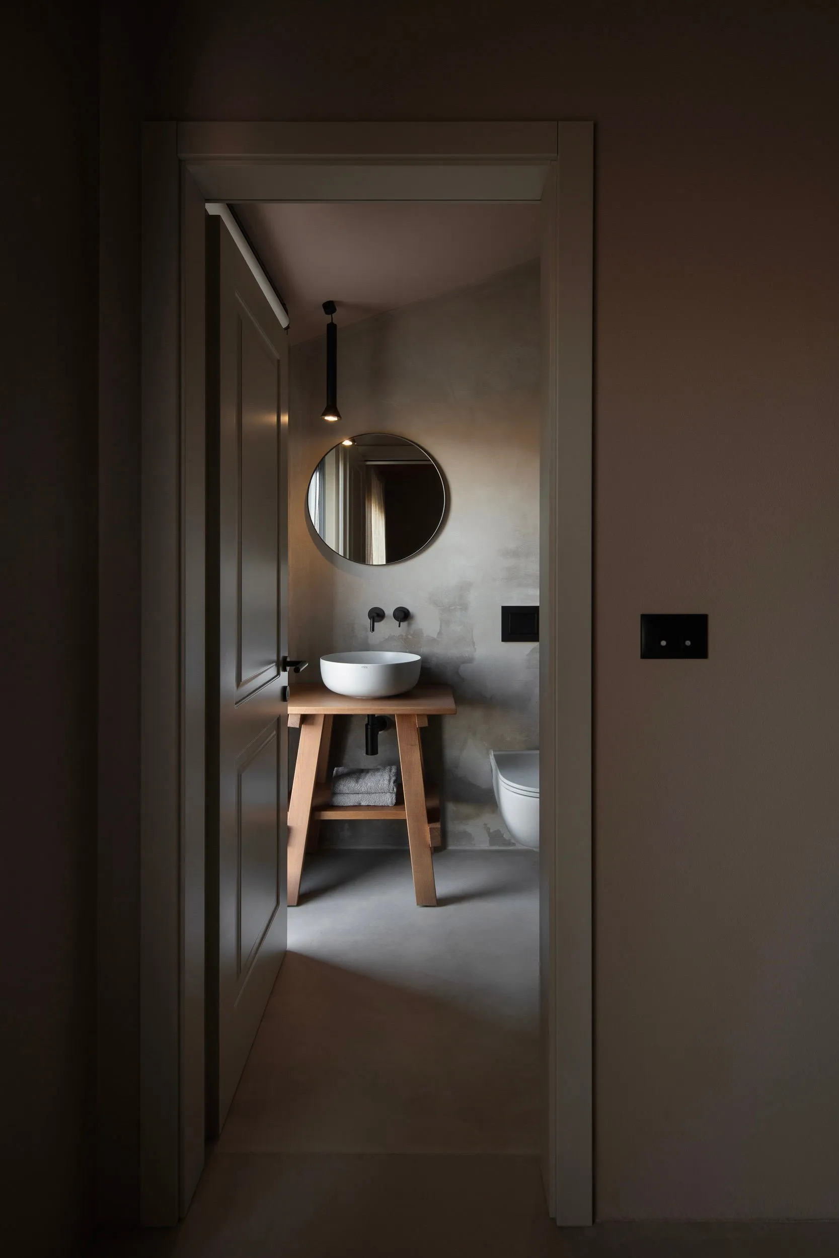 A minimalist bathroom with concrete walls, a circular mirror, and a white sink on a wooden stand.