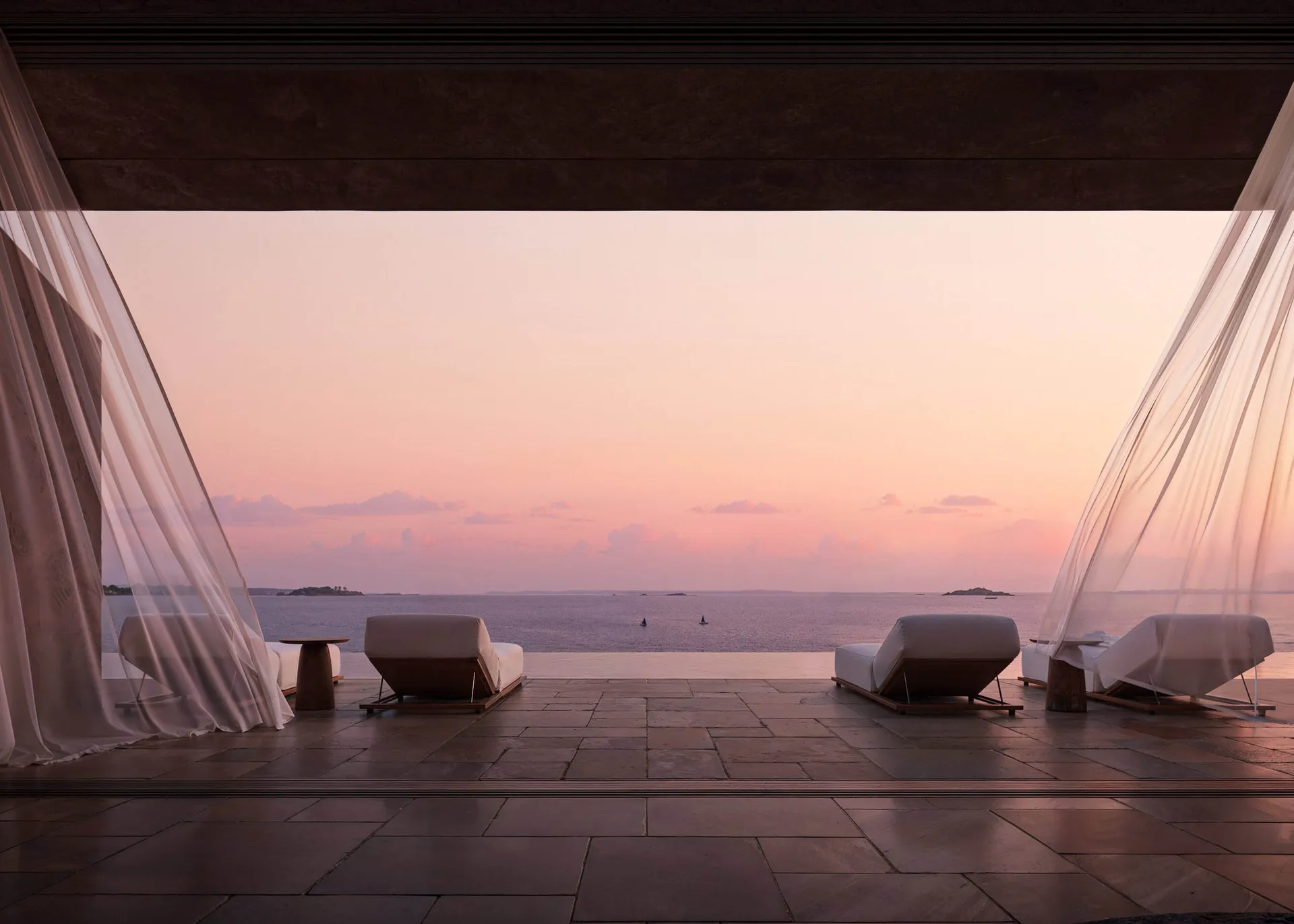 A minimalist outdoor lounge featuring white fabric seating and wooden frames, overlooking the ocean at sunset.