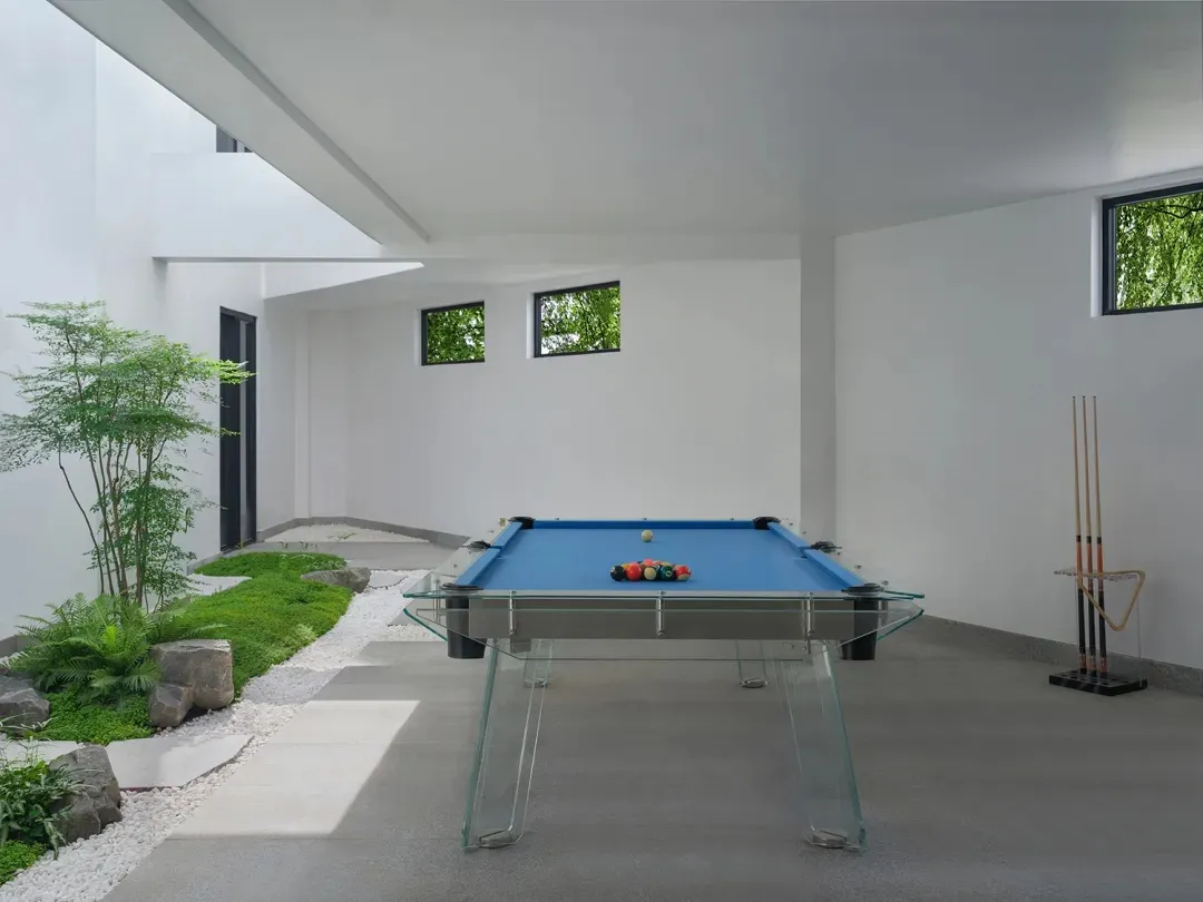 A modern living room with a glass pool table, concrete floor, and greenery.