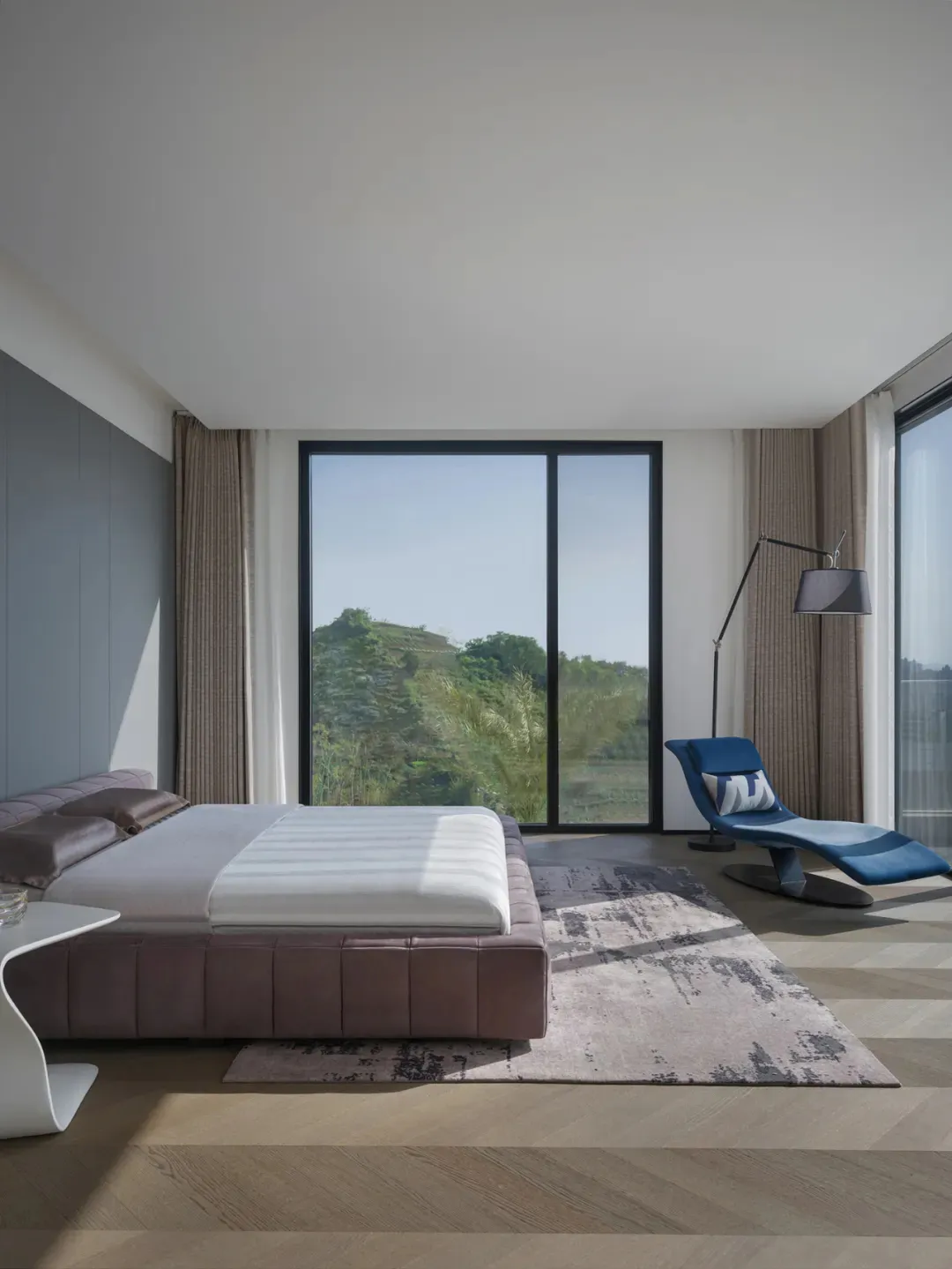 A modern minimalist bedroom featuring a sleek, upholstered bed with a glass headboard, a smart touch bedside table, and a con