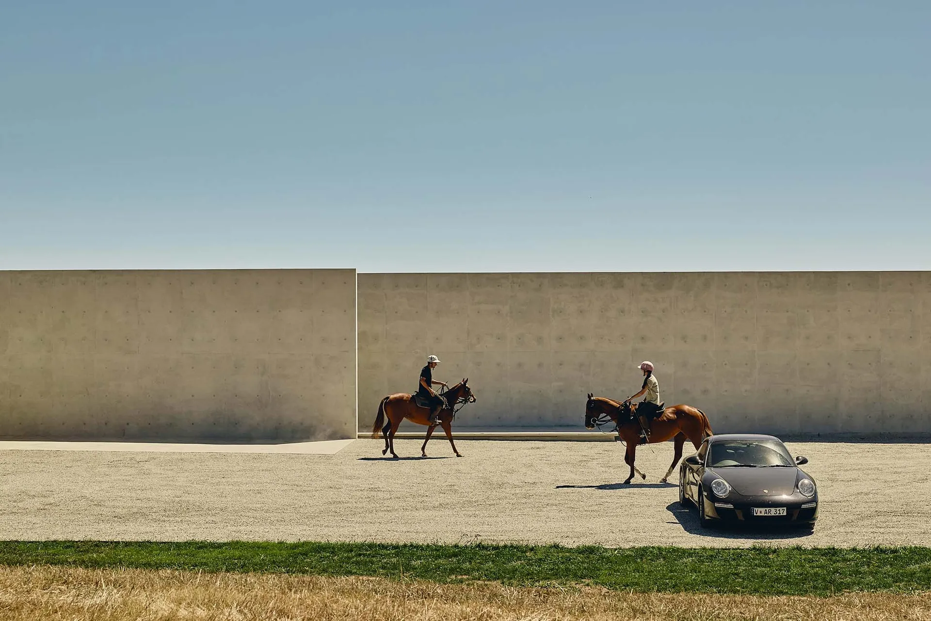 Two people riding horses in front of a concrete wall, with a car parked nearby.