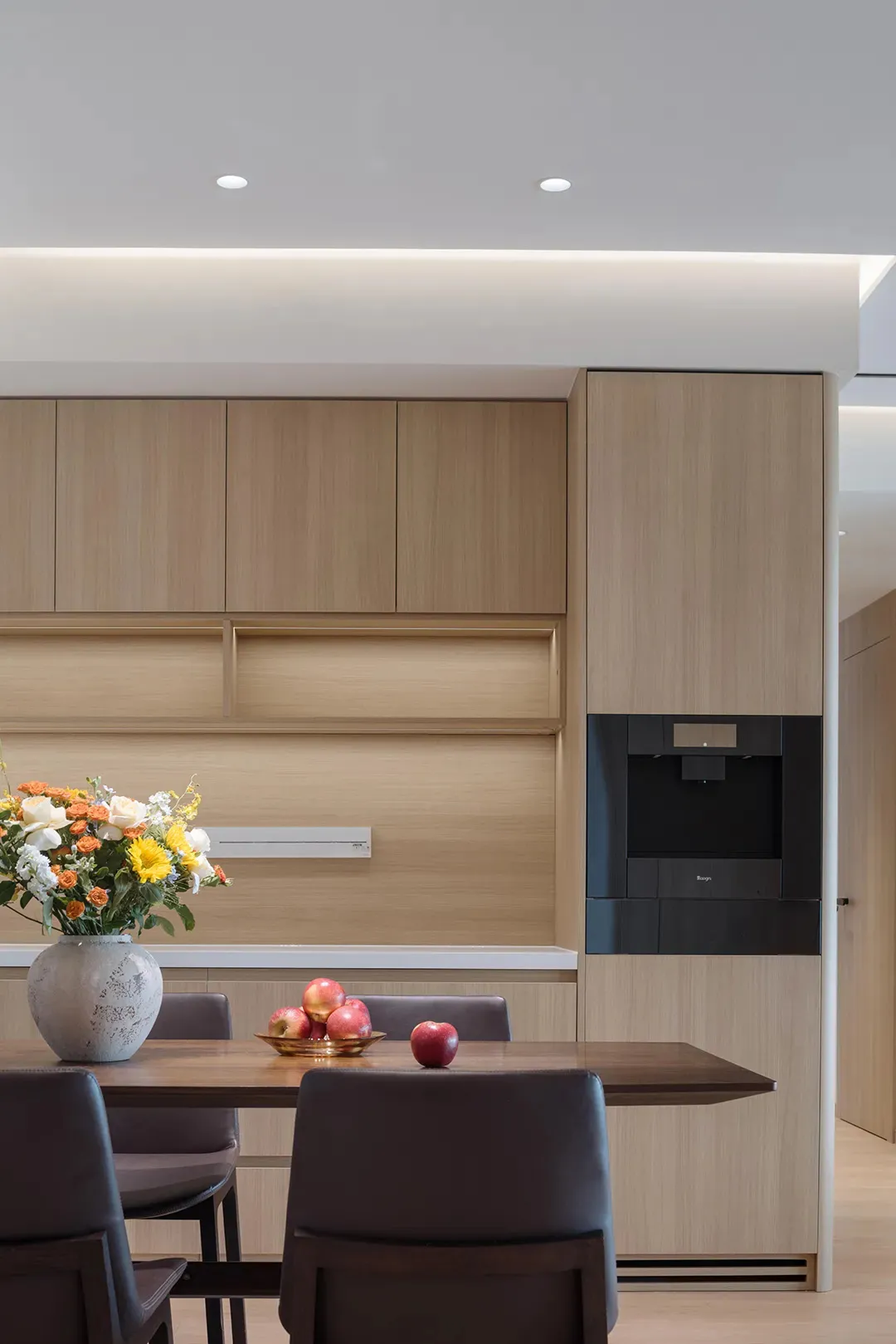 Modern residential kitchen featuring timber cabinetry, a dining table with fruits and a vase of flowers.