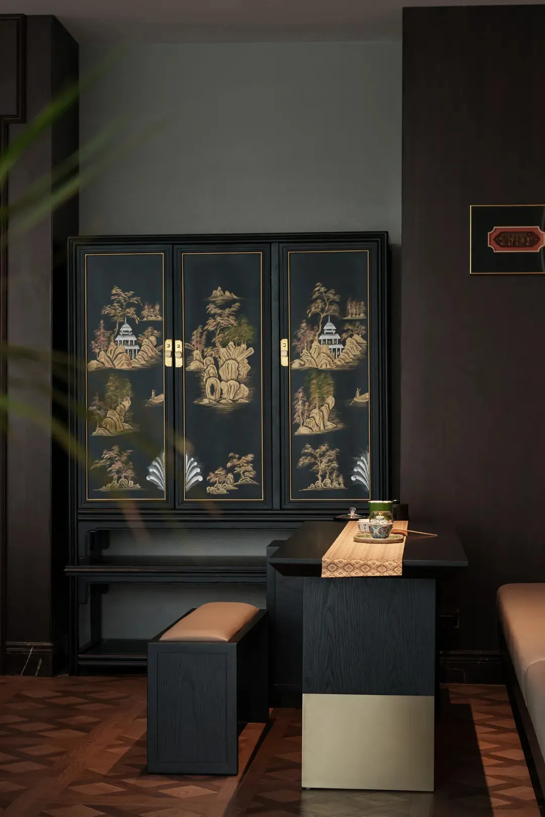 A black lacquer cabinet with gold inlay depicting Chinese landscapes, placed in a darkly painted room with a low table in fro
