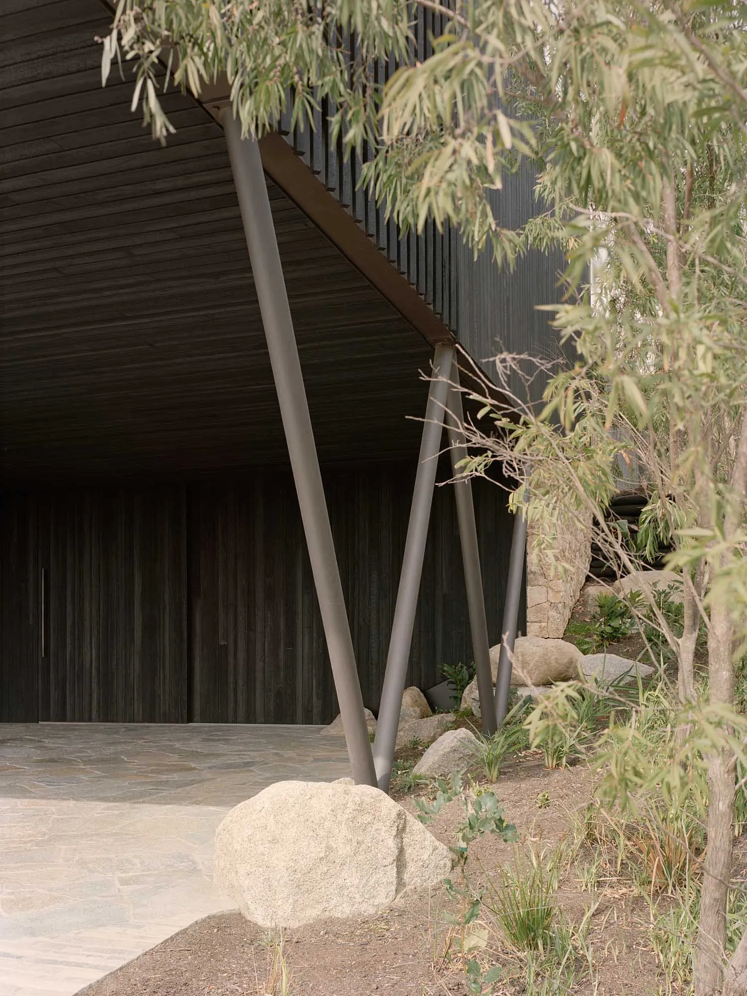 Outdoor minimalist entryway with wooden ceiling and metal frame supported by wooden pillars and surrounded by greenery on a s