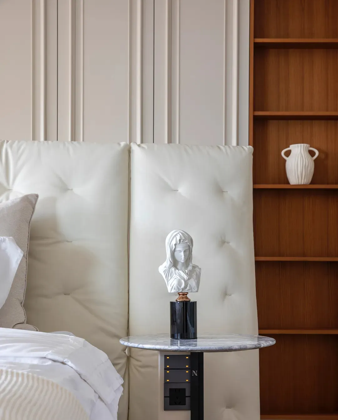 A minimalist bedroom featuring a leather headboard with tufted cushions, a marble bedside table, and a wooden bookshelf with