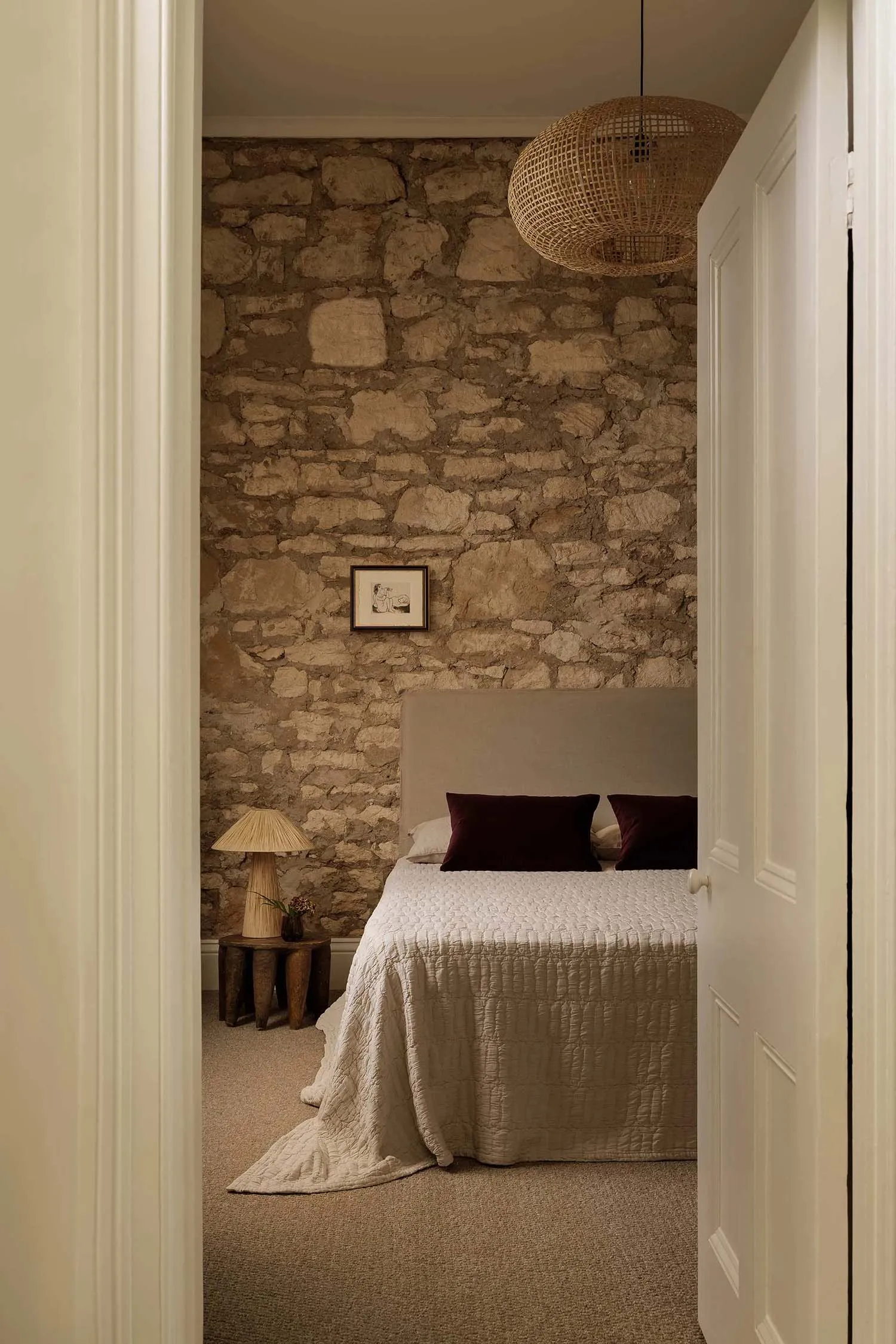 Minimalist bedroom featuring a stone wall, beige carpet, bed with burgundy pillows, wooden stool with lamp, and a woven penda