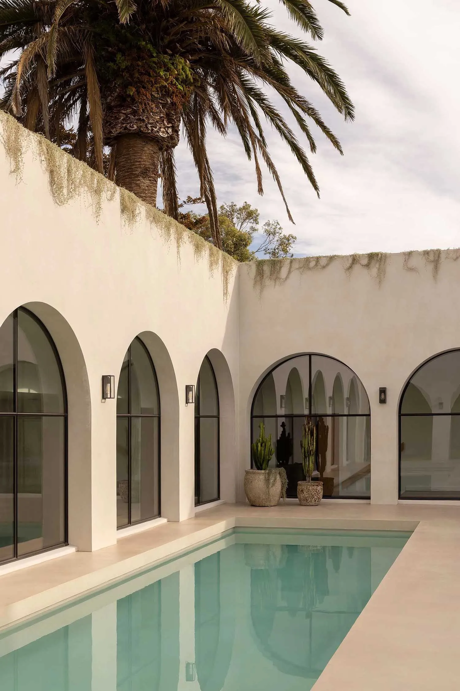 Modern house with white plaster exterior, arched windows, and a swimming pool surrounded by palm trees.