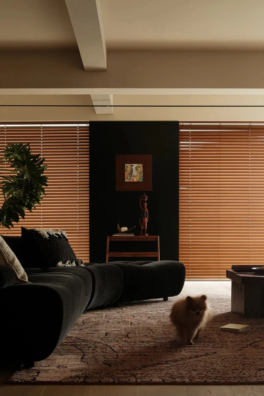 A modern minimalist living room with wooden blinds, a dark-colored sofa, and a small dog walking on a patterned rug.