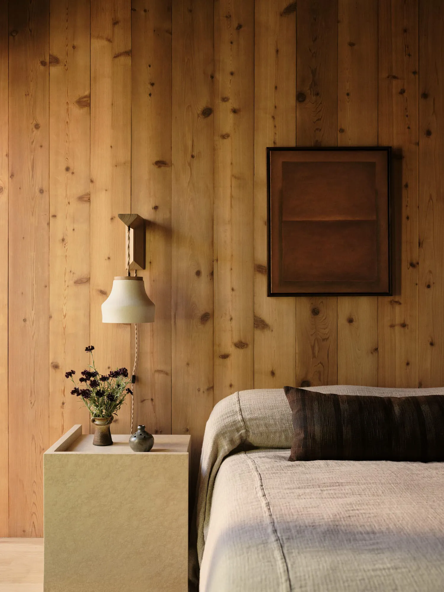 A bedroom with wooden walls, a bed, and a side table with a vase of flowers and a small lamp.
