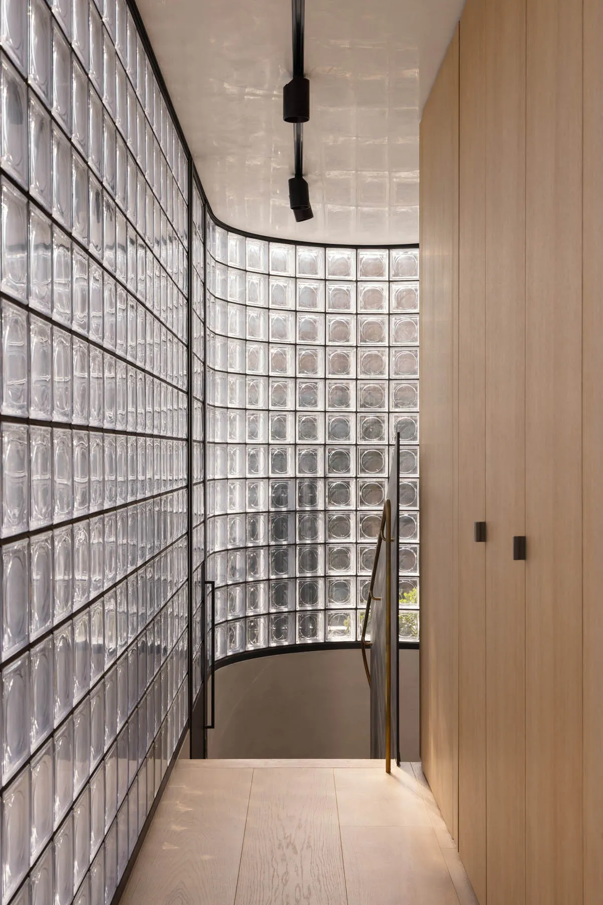 A modern staircase with transparent glass blocks leading downward, bordered by wooden panels and illuminated by overhead ligh