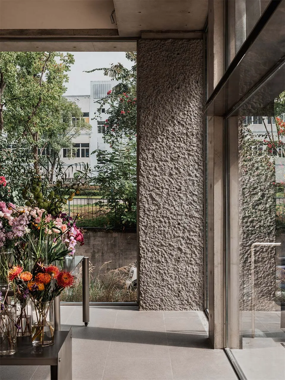 Modern lobby with concrete walls and large windows, featuring a vase of flowers and greenery.