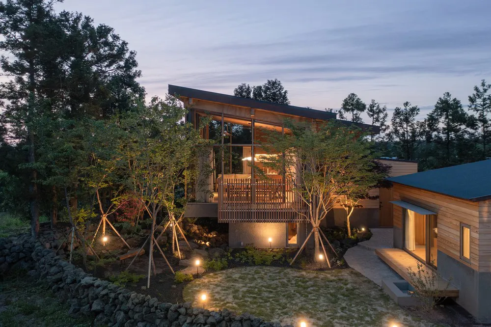 A serene modern residential house with stone accents, wooden exterior, and glass windows against a twilight sky.