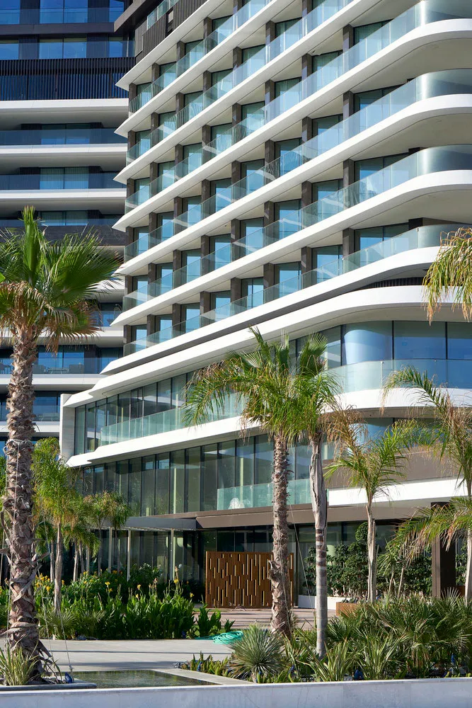 Modern residential building with glass windows and concrete balconies, surrounded by palm trees and greenery.