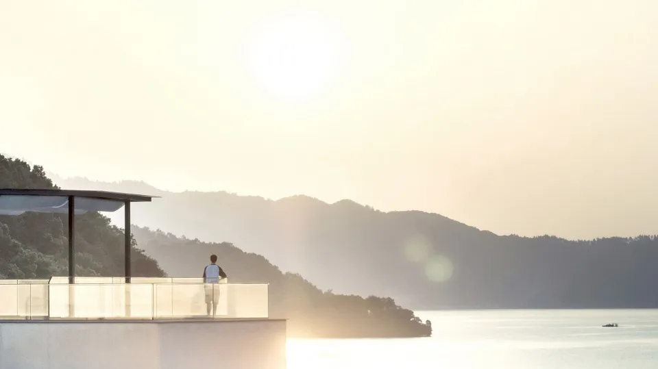 Person standing on a balcony overlooking a serene lake surrounded by mountains during sunset.