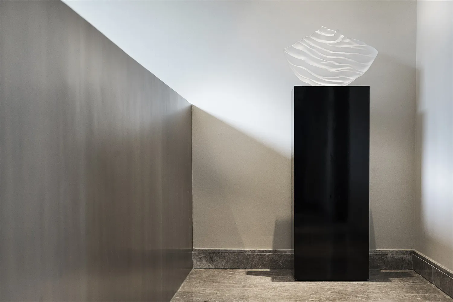 A modern, abstract glass sculpture with a wave-like design displayed on a black pedestal in a minimalist interior setting.
