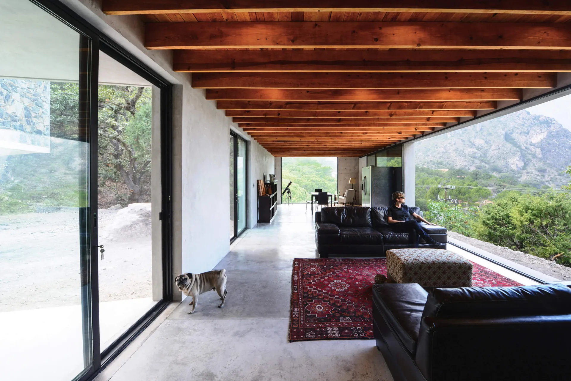 A modern living room with concrete flooring, black sofas, and a wooden ceiling with exposed beams.