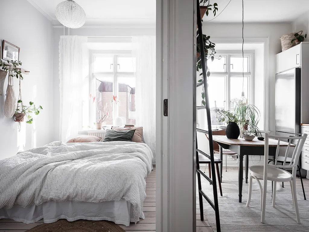 A minimalist bedroom featuring a cozy bed covered in light-colored cotton textiles, surrounded by natural light and green pla