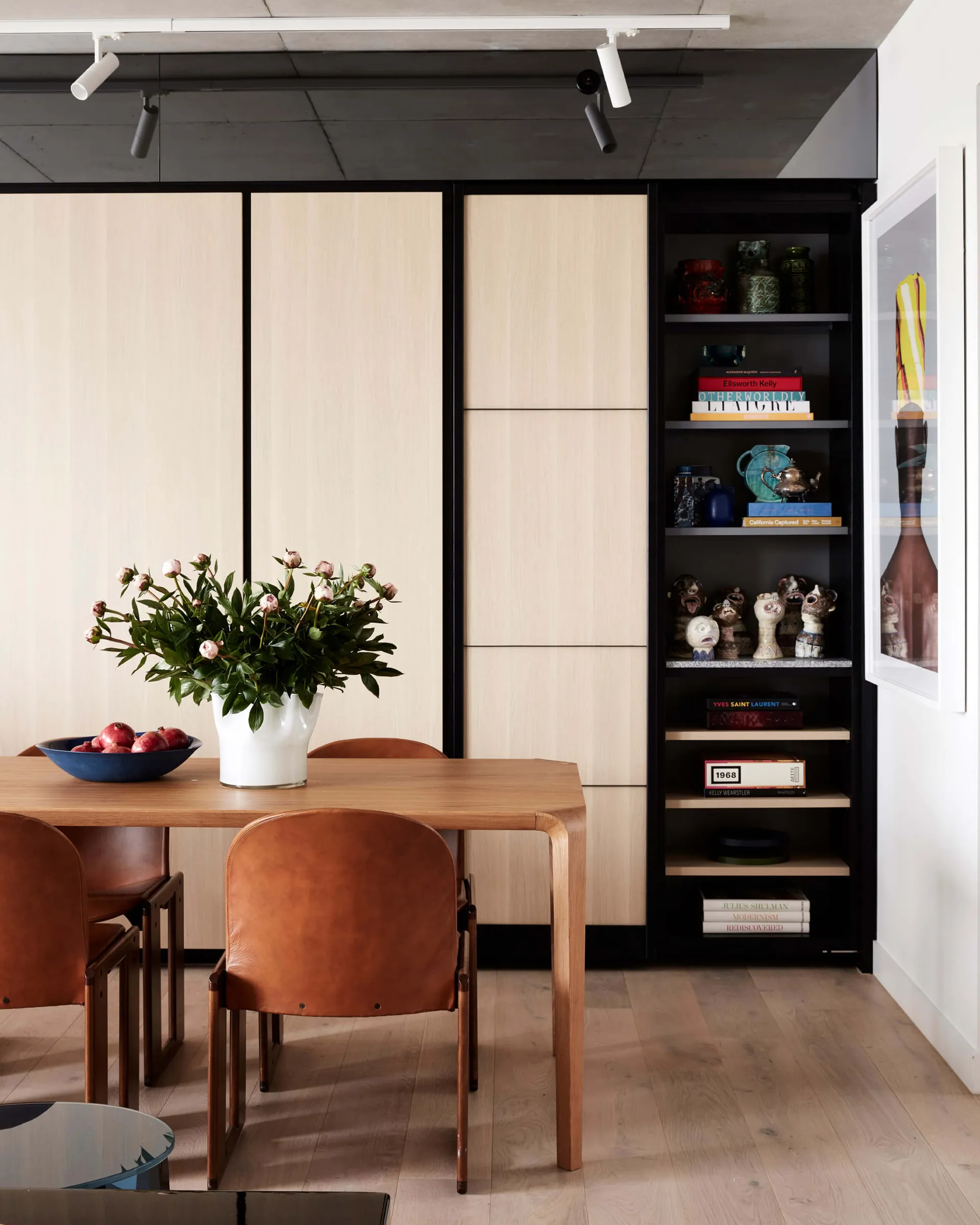 Contemporary dining room featuring a wooden table, glass vase with flowers, and a bookshelf displaying decorative items.