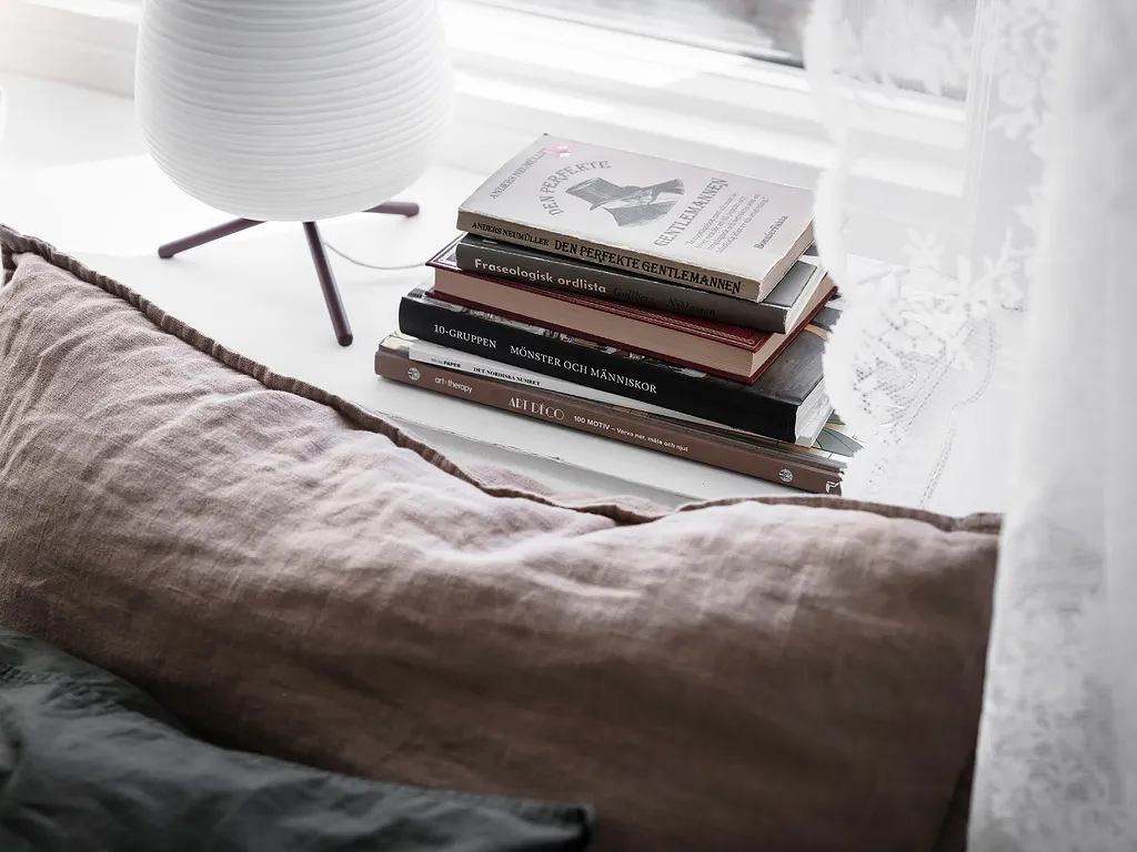 Stack of books on a white surface with a white table lamp and curtains in the background, featuring a brown pillow and cotton