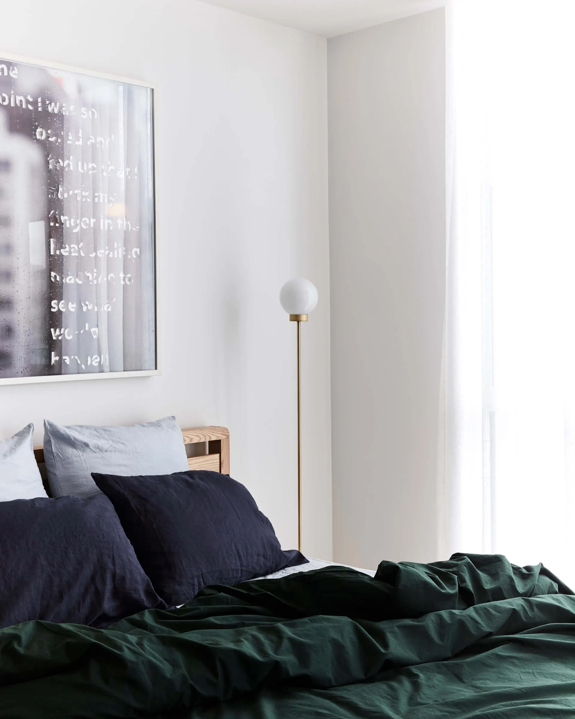 A minimalist bedroom with white walls, white flooring, and white blinds, featuring a bed with dark blue and green bedding, wh