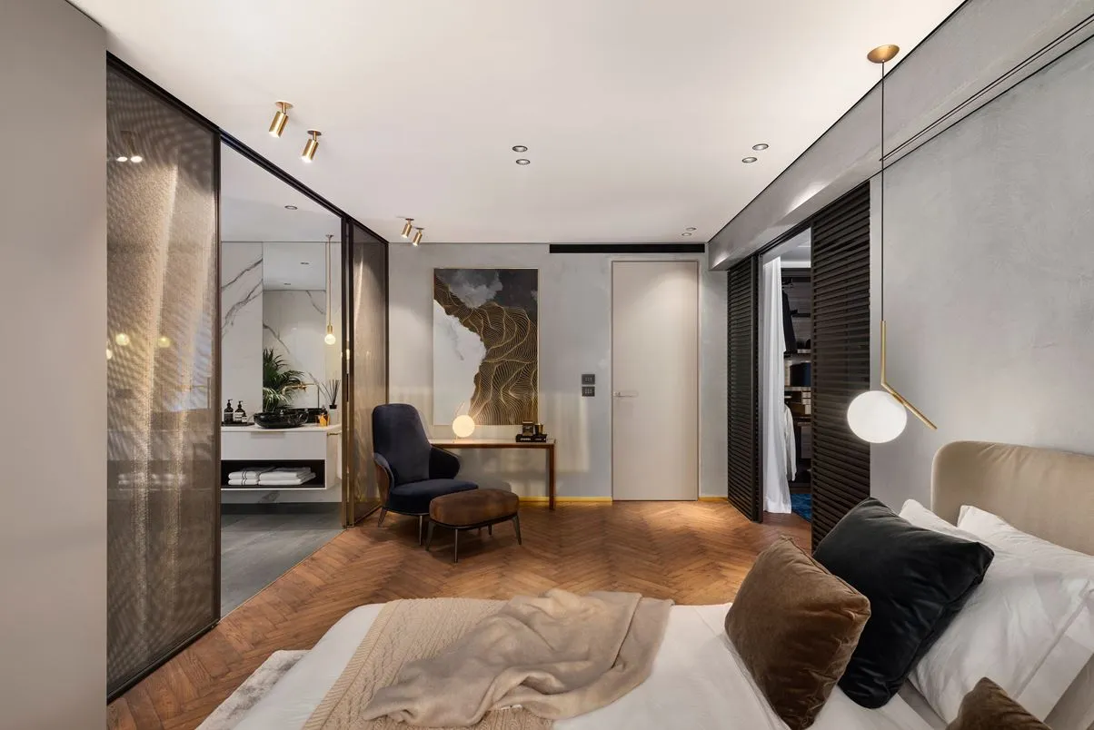 Modern bedroom with wooden flooring, a bed with a beige blanket and black pillows, and a chair in the corner of the room.