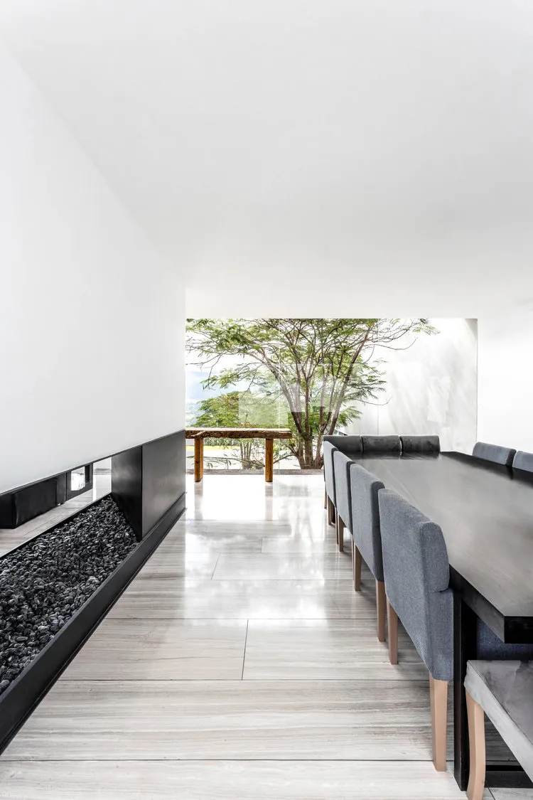 A modern minimalist dining room featuring concrete flooring and walls, with a long dining table, grey upholstered chairs, and