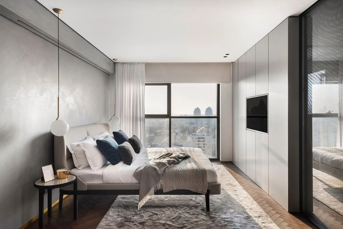 Modern minimalist bedroom with a large bed, white and gray pillows, and a view of the city through the window.