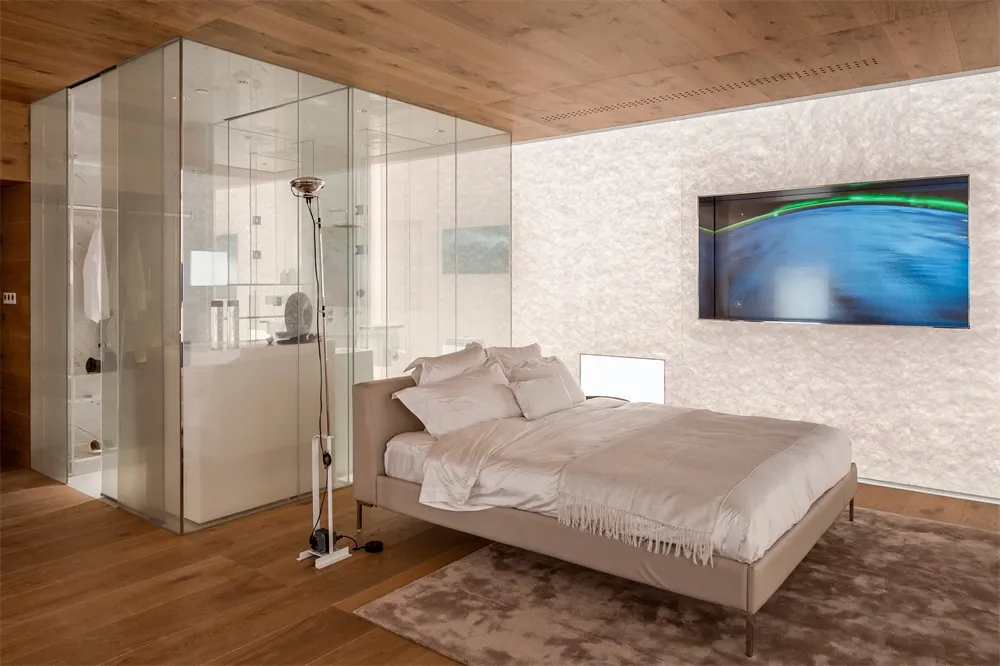 Modern minimalistic bedroom with a beige bed, glass enclosed bathroom, wooden flooring, and a flat-screen television.