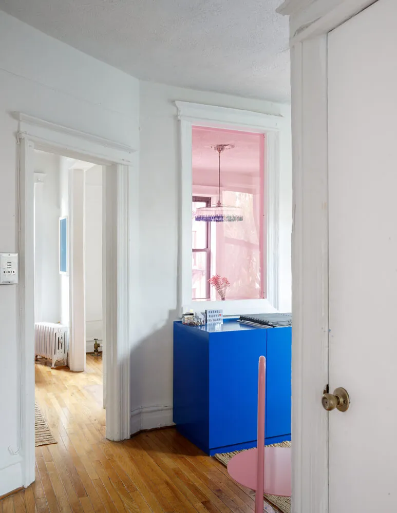 A home office space with white walls, a bright pink wall, a blue table, and a pink chair.