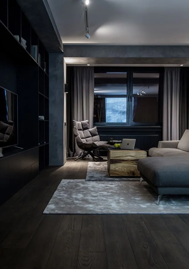 A modern living room with dark wooden floors, a cozy grey sofa, and a stylish armchair, featuring a large bookshelf, a wooden