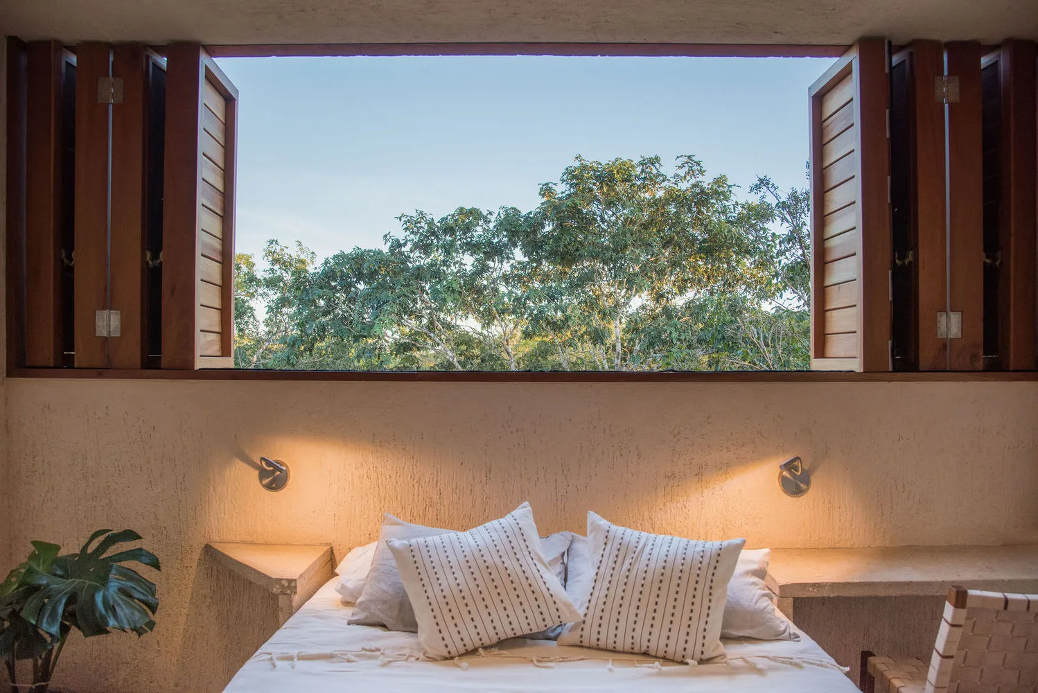 A cozy, modern bedroom with a large window overlooking lush greenery, illuminated by warm sconces.