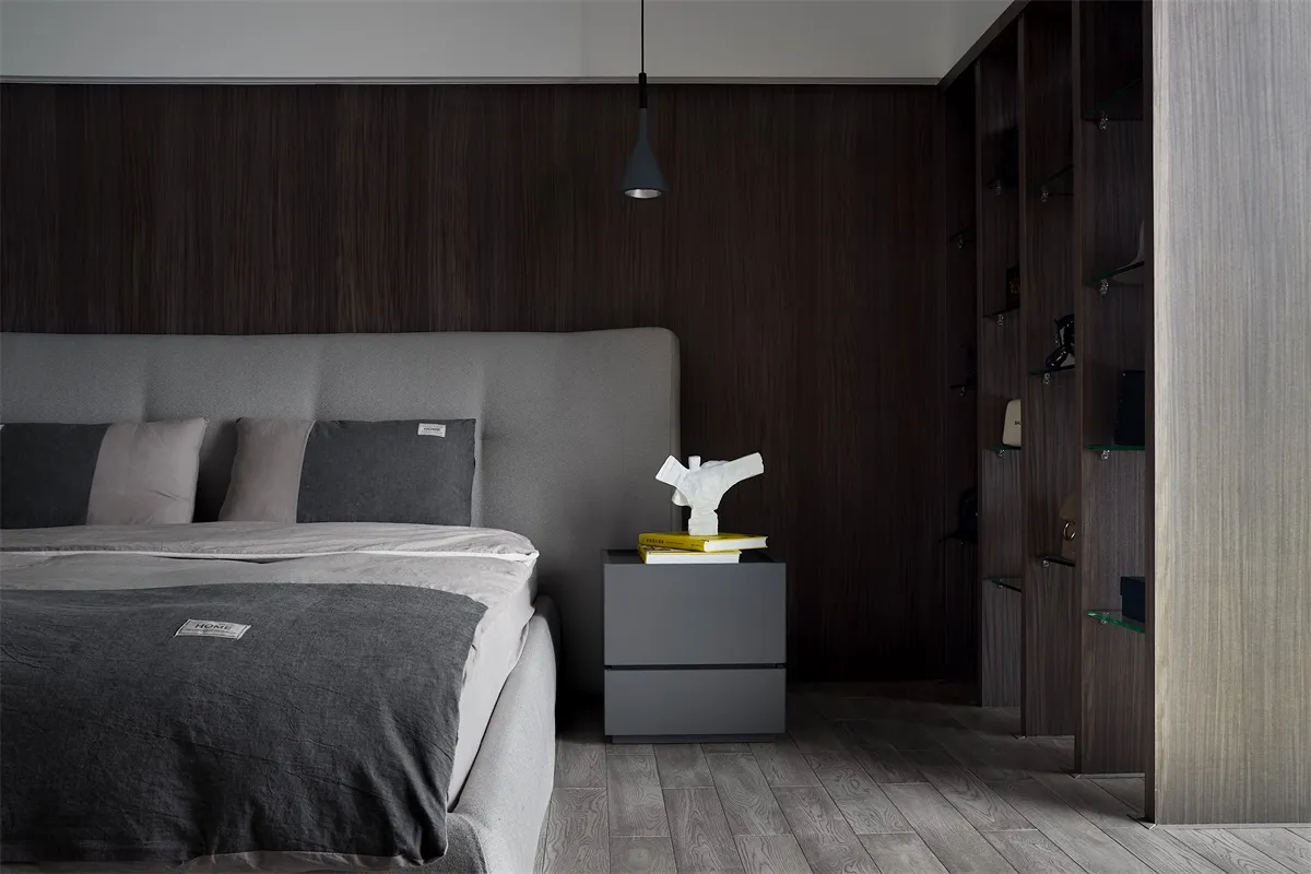 A modern minimalist bedroom featuring wood-paneled walls, a grey upholstered bed, and a grey wooden nightstand with books and