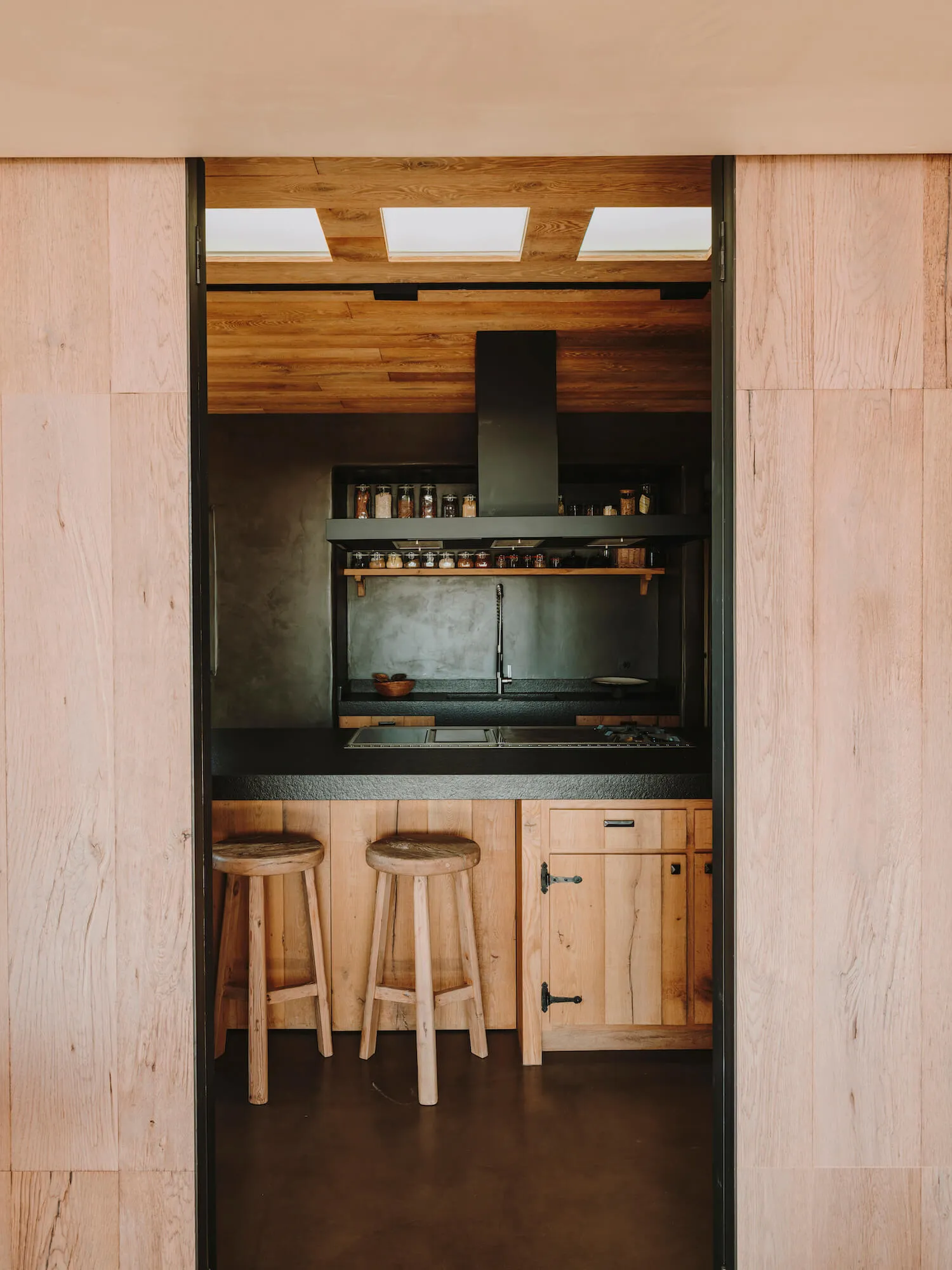Modern kitchen with wooden ceiling and floor, featuring a black countertop, wooden stools, and a minimalist design.