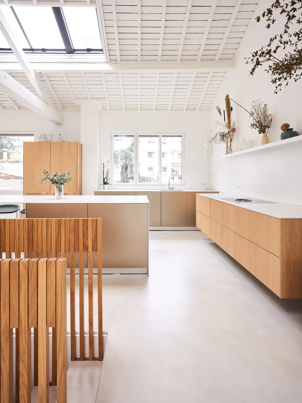 Modern kitchen features wooden cabinets and white flooring, with a minimalist design and large windows.