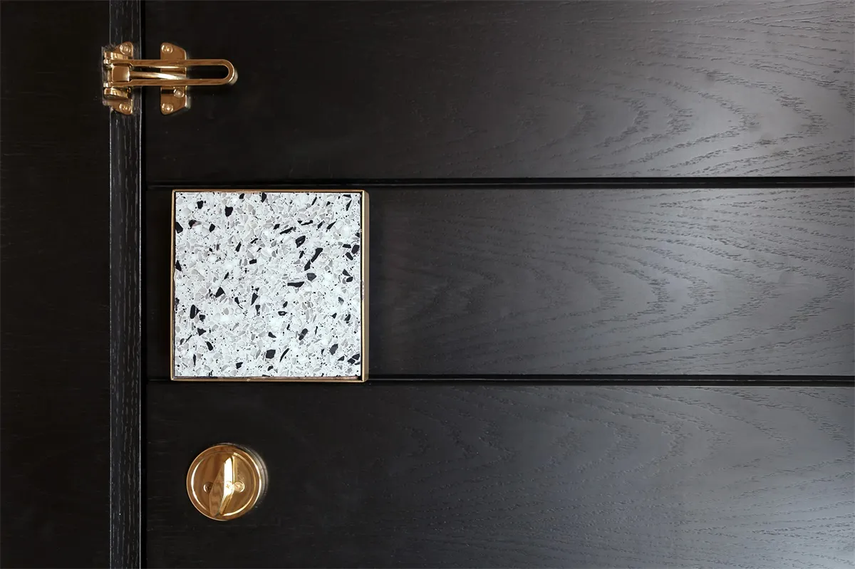 An elegant, black-colored wooden door with a geometric, white speckled terrazzo tile and a gold latch.