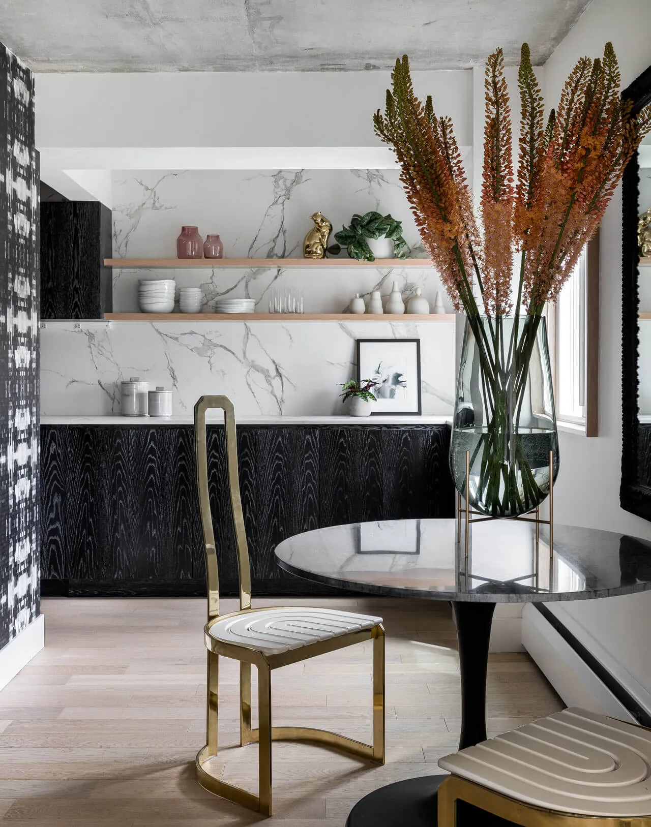 Contemporary dining area with marble countertops, glass vase with orange flowers, and black and gold metallic chairs.