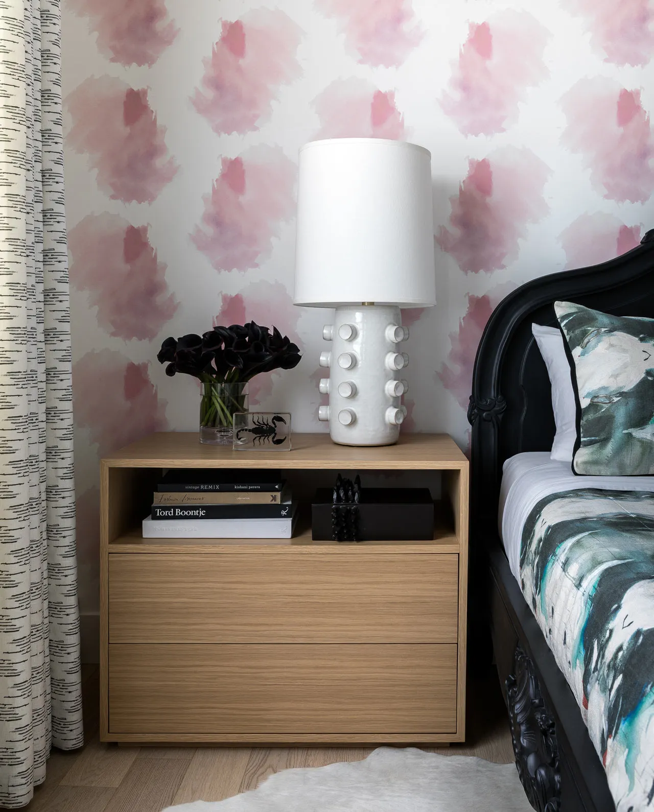 A minimalist bedroom featuring a black bedside table with a white lamp, black flowers, and books, placed against a wall with