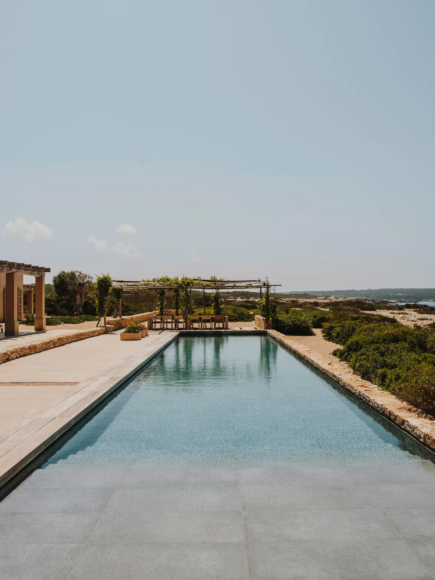 A minimalist outdoor space featuring a long, rectangular swimming pool bordered by concrete surfaces, surrounded by greenery